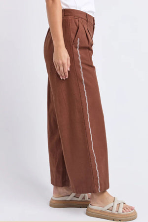A person stands sideways against a plain background wearing the relaxed-fit, high-waisted Foxwood Paradise Pant featuring vertical white wavy stitches, paired with a white sleeveless top and beige sandals.