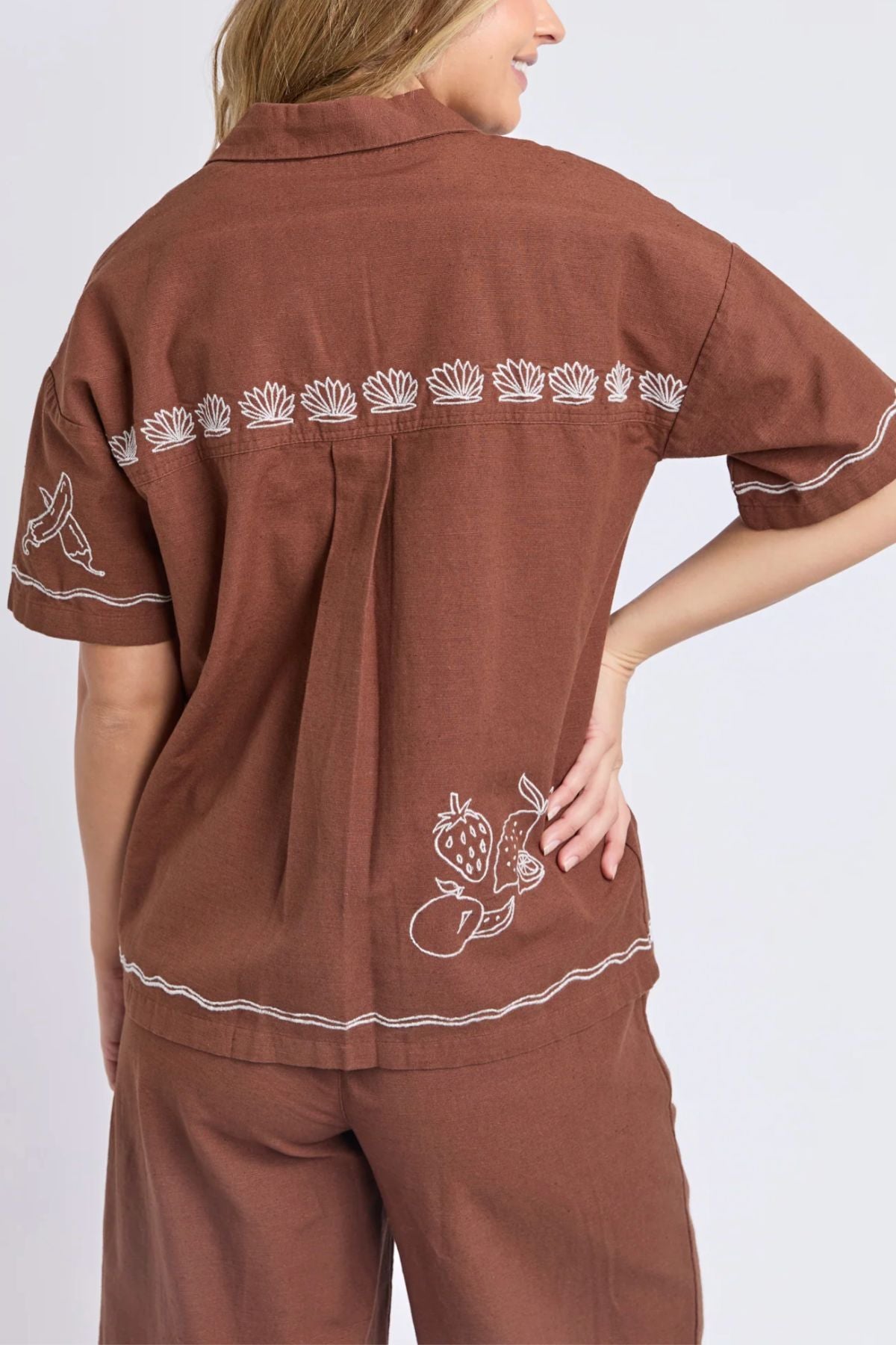 A woman wears the Foxwood Paradise Shirt, a relaxed-fit top featuring white embroidered fruit, flower, and bird designs on the back and sleeves. She stands with one hand on her hip and pairs it with matching pants.