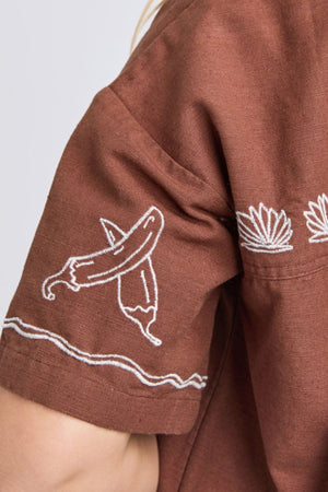Close-up of the Foxwood Paradise Shirt, a relaxed-fit brown shirt featuring white embroideryโa banana on the sleeve and abstract leaves on the upper back and shoulder seam.