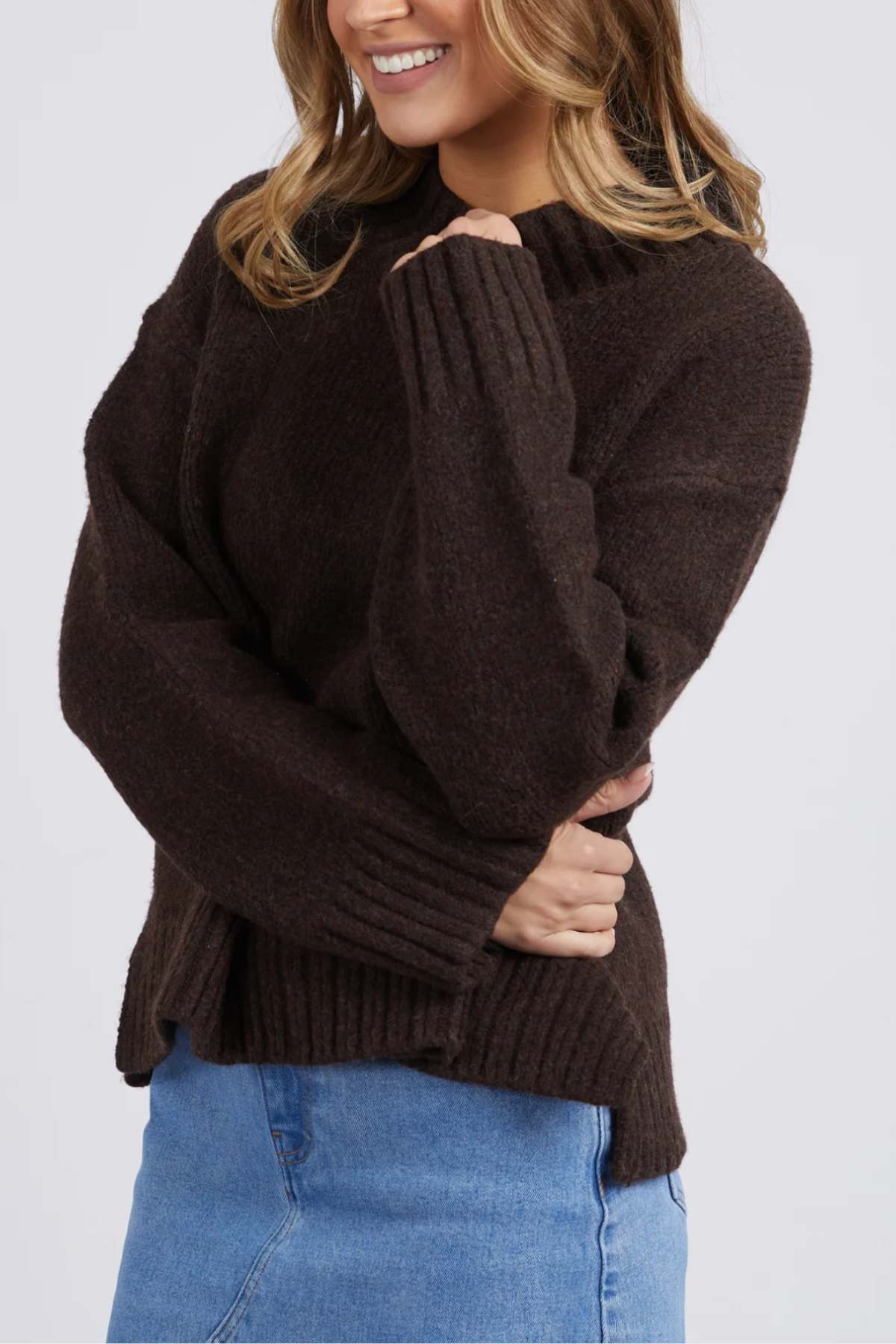 A woman smiles, wearing the Foxwood Pepper Knit—a cozy, dark brown long-sleeve sweater—paired with a light blue denim skirt. With her left arm crossed over her body, only the lower part of her face is shown.