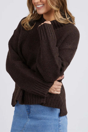 A woman smiles, wearing the Foxwood Pepper Knit—a cozy, dark brown long-sleeve sweater—paired with a light blue denim skirt. With her left arm crossed over her body, only the lower part of her face is shown.