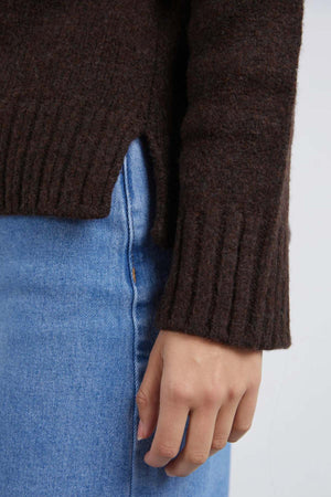 A close-up of a hand resting by the side, wearing the Foxwood Pepper Knit—a relaxed fit brown sweater with a side slit—paired with blue denim jeans against a plain, light background. Ideal for casual looks.