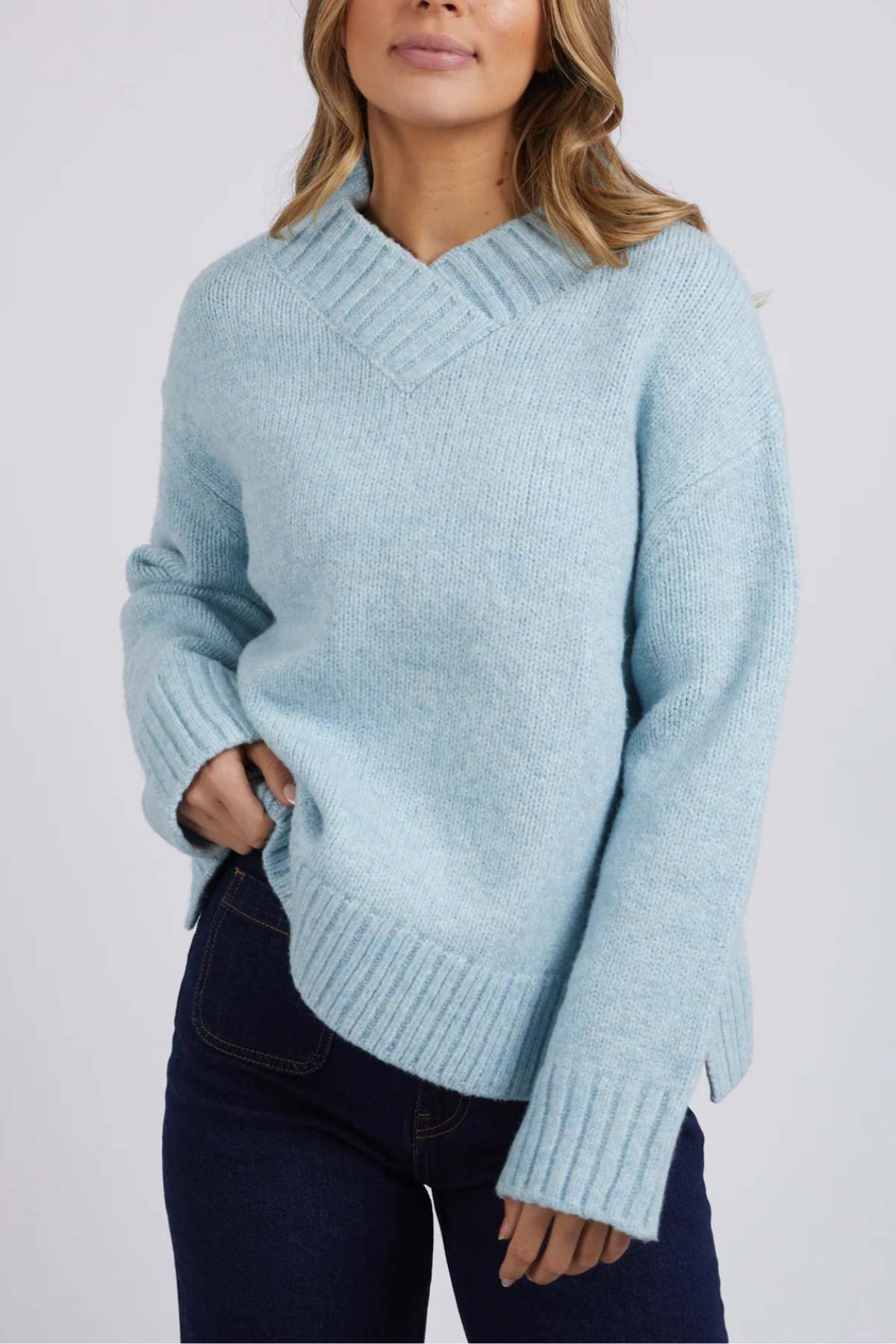 Wearing the light blue Foxwood Pepper Knit sweater with a cozy V-neck and relaxed fit, the person stands against a plain background. Their medium-length wavy hair pairs well with dark blue jeans, as they casually place one hand in their pocket, exuding effortless style.