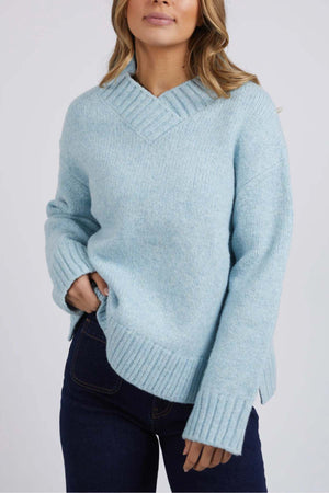 Wearing the light blue Foxwood Pepper Knit sweater with a cozy V-neck and relaxed fit, the person stands against a plain background. Their medium-length wavy hair pairs well with dark blue jeans, as they casually place one hand in their pocket, exuding effortless style.