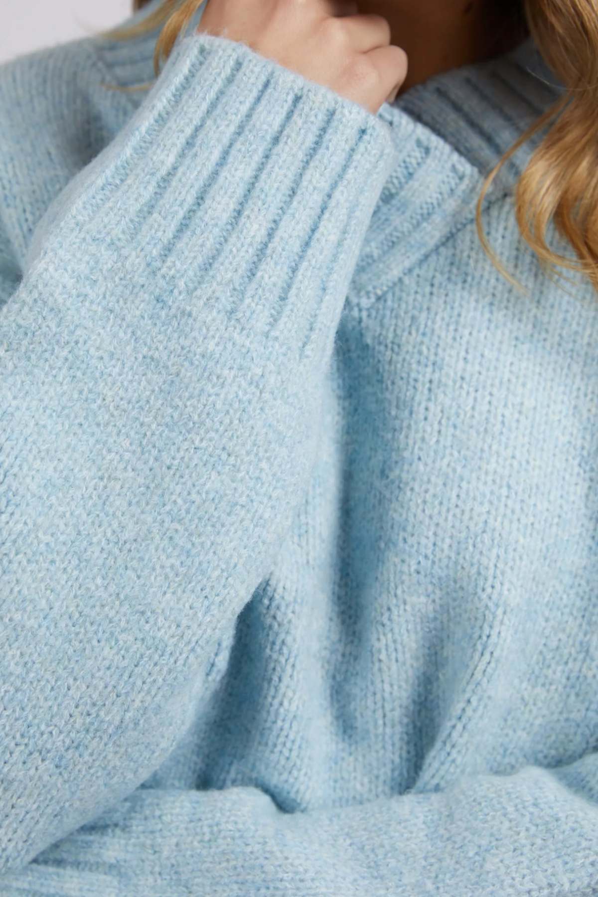 A person is wearing the Foxwood Pepper Knit, a light blue chunky knit sweater with ribbed cuffs and a thick collar. The cozy blend has a relaxed fit, with long hair resting on their chin, partially covered by the sleeve.
