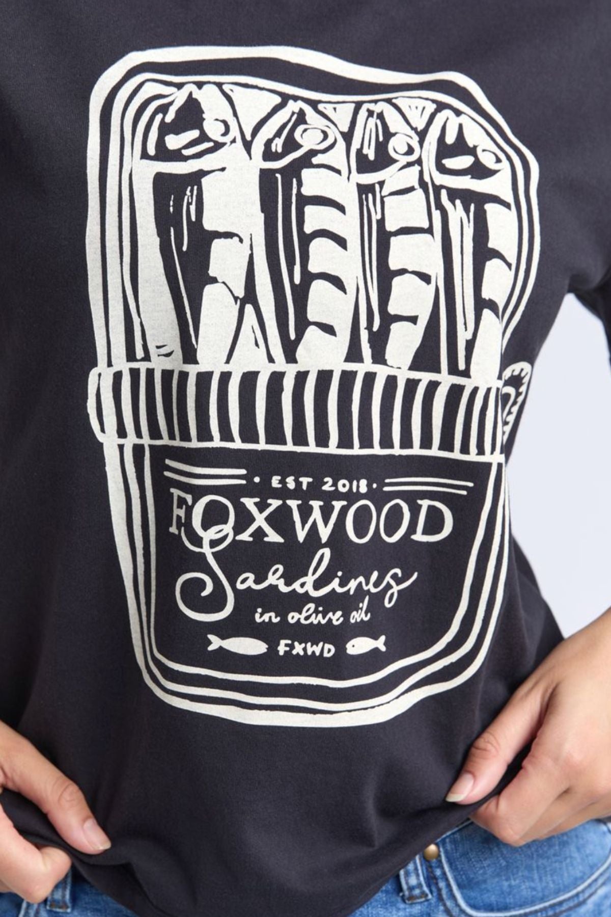 Wearing the Foxwood Pesce Tee—a 100% cotton, coastal-inspired shirt—features four sardines in a tin can and "Foxwood Sardines in olive oil EST 2018 FXWD" below the illustration.