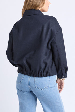 A woman with light brown hair wears the Foxwood Pippa Jacket, a dark navy wool blend with an elastic waistband, paired with light blue high-waisted jeans against a plain white backgroundβan ideal cosy outerwear choice.