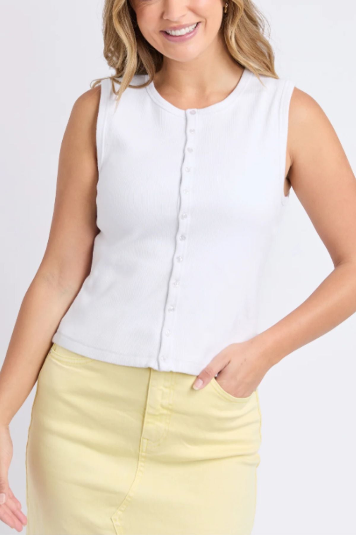 A woman smiles against a plain light background, wearing the Foxwood Raf Button Through Tank in sleeveless white and a pale yellow skirt, standing with one hand in her pocket.