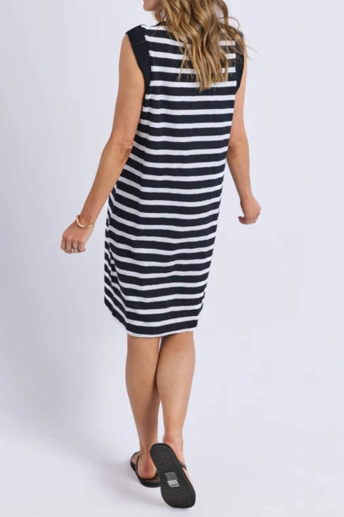 A woman with wavy, light brown hair is seen from behind wearing a sleeveless, knee-length Foxwood Reform Stripe Dress with black and white stripes and black slip-on shoes, walking against a plain light background.