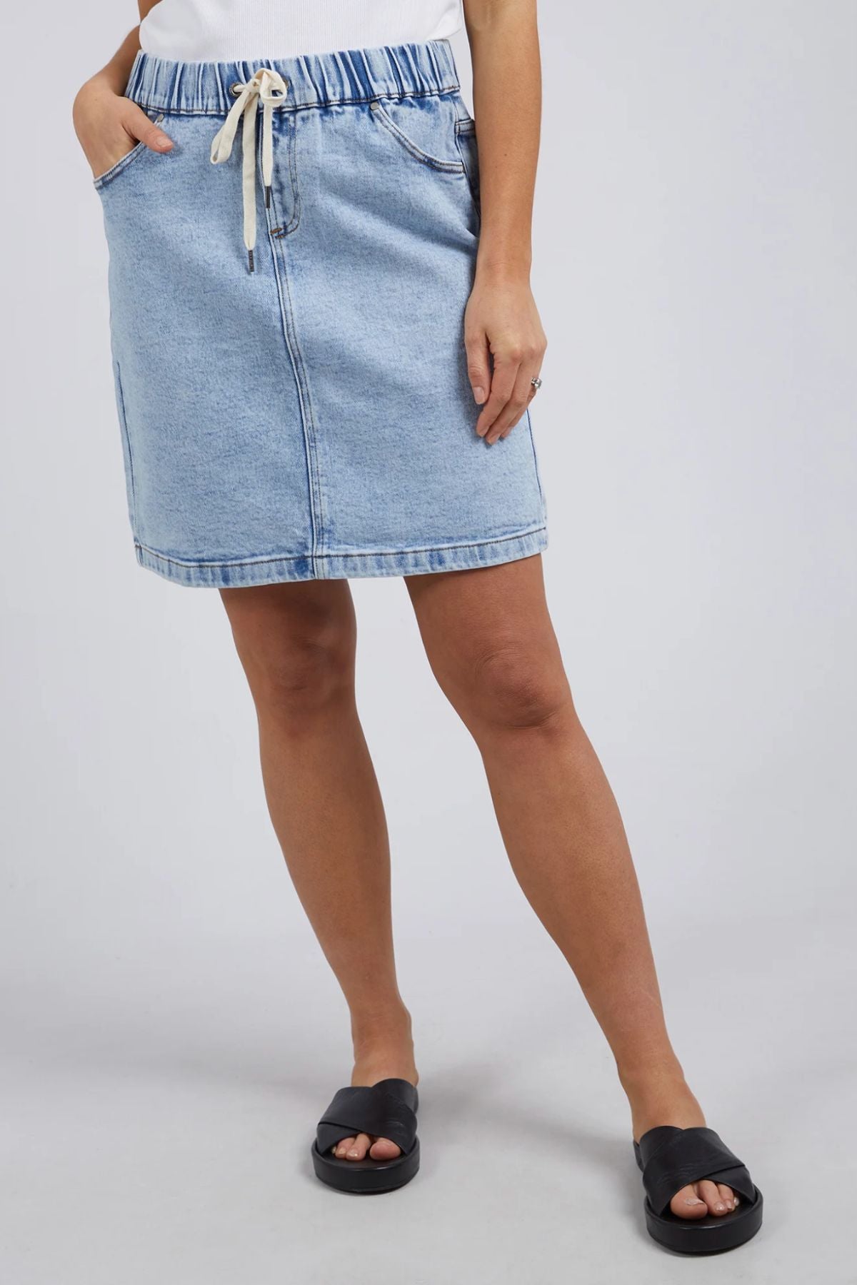 A person in a light blue Rio Skirt with side pockets and a white drawstring waist, styled with a white top and black slide sandals, stands casually against a plain gray background, hand tucked into the pocket.