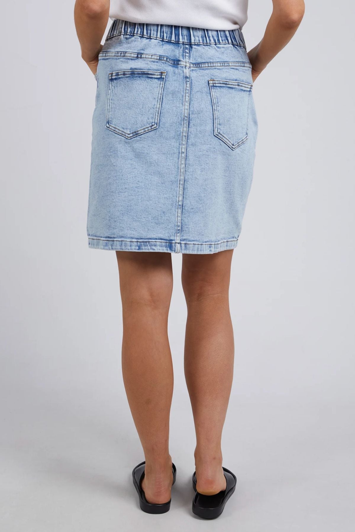 A person in a Foxwood Rio Skirt, light blue with two back pockets and visible stitching for a stretchy fit, paired with black slip-on sandals, stands facing away in front of a neutral background.