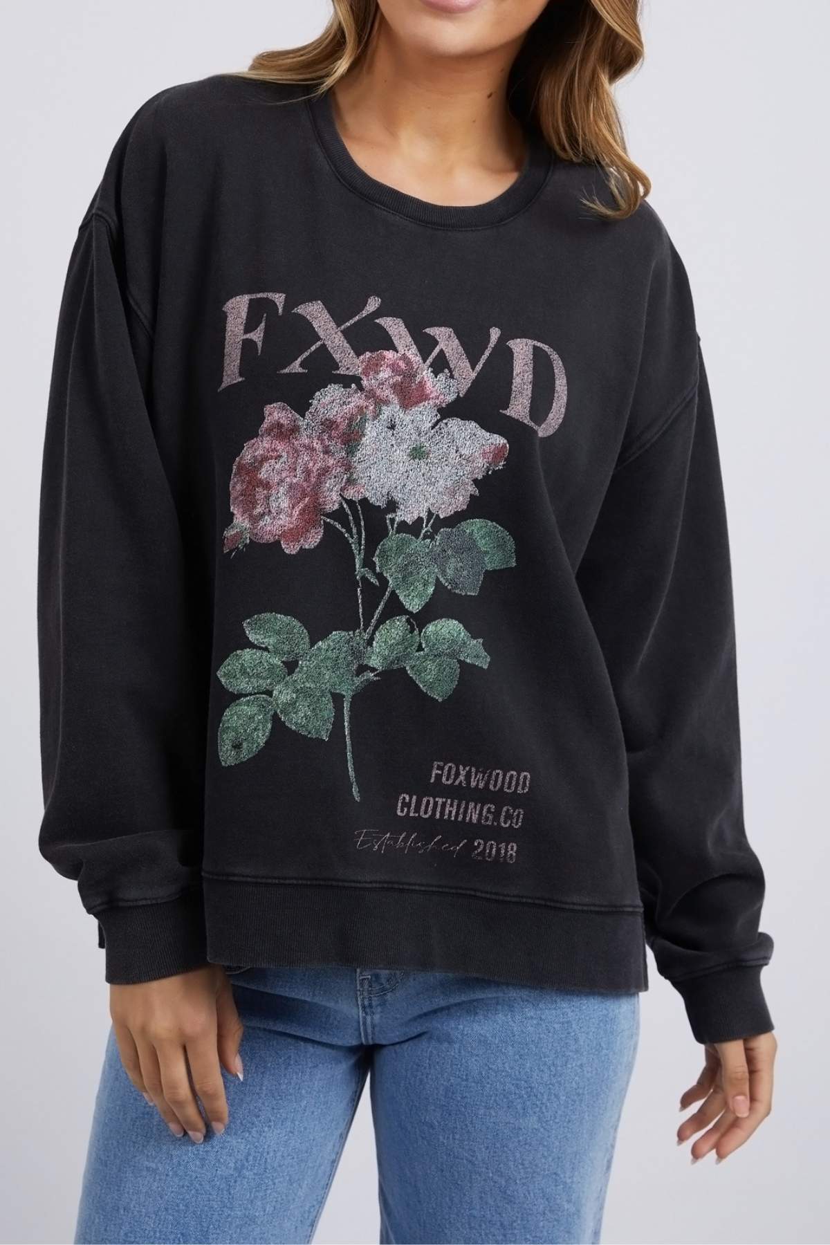 A person wears the black Foxwood Rosa Crew sweatshirt, 100% cotton, featuring a "FXWD" chest print with a pink and green floral design, and "Foxwood Clothing Co Established 2018" below. Blue jeans complete the outfit.