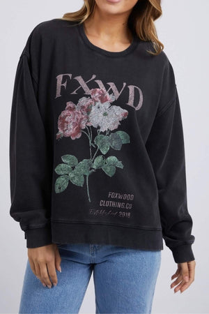 A person wears the black Foxwood Rosa Crew sweatshirt, 100% cotton, featuring a "FXWD" chest print with a pink and green floral design, and "Foxwood Clothing Co Established 2018" below. Blue jeans complete the outfit.