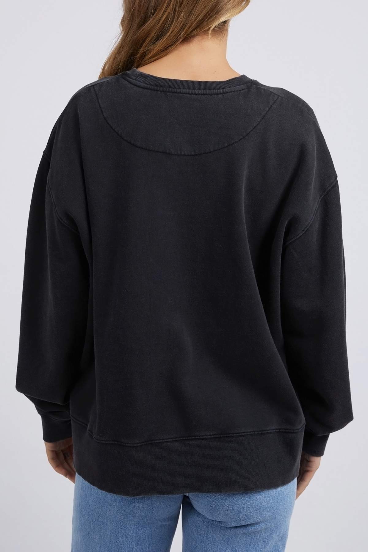 Facing away, a person with long hair wears a loose-fitting Foxwood Rosa Crew sweatshirt made of 100% cotton and light blue jeans, with subtle chest print details. The background is plain and light-colored.