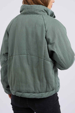 A person is shown from the back wearing the Foxwood Rosalee Denim Jacket in sage green with a cozy cotton denim feel. The high collar pairs well with black jeans, and their brown hair is visible while their face is turned away.