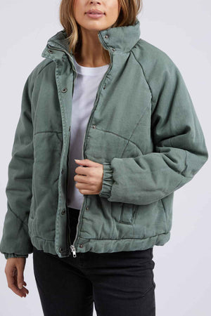A person wearing the Foxwood Rosalee Denim Jacket, a cozy green cropped slouchy cotton denim, partially unzipped over a white t-shirt and black pants, stands against a plain light background holding one side of the jacket.