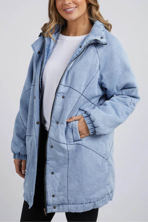 A woman in the Foxwood Rosalee Longline Jacket, vintage washed denim and 100% cotton, pairs it with a white tee and black pants, smiling with one hand in her pocket against a plain background.