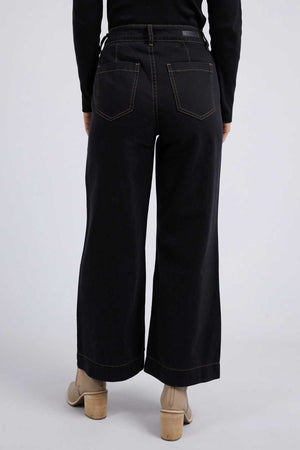 A person is seen from the rear wearing Foxwood Royal Wide Leg Jeans, characterized by high-waisted, wide-leg black denim with contrast stitching. They complement the look with beige heeled ankle boots and a black top against a plain light gray background.