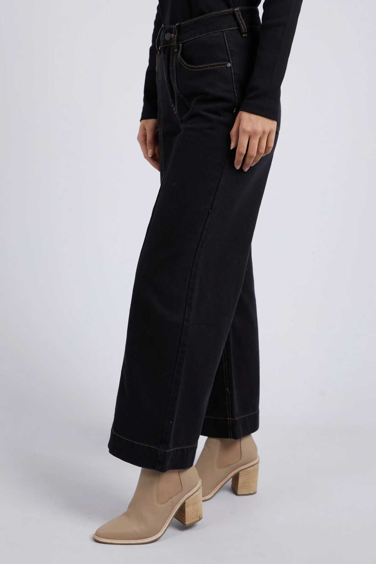 Striking a pose, a person wears Foxwood Royal Wide Leg Jeans in black with contrast stitching paired with a sleek black top. Their light beige ankle boots featuring chunky heels stand out against the plain, light-colored background.