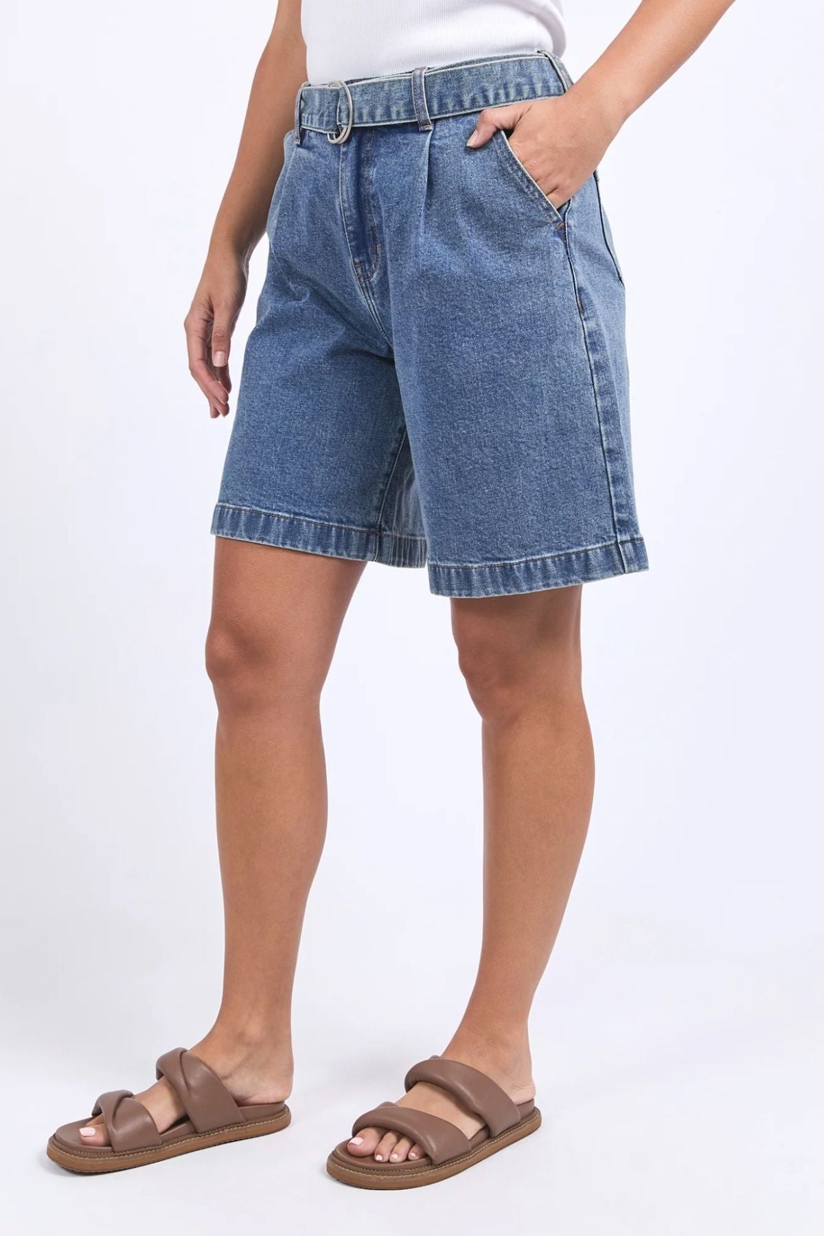 Someone stands against a plain white background wearing the Foxwood Saint Shortβknee-length, stretchy denim shorts with a beltβpaired with brown sandals.