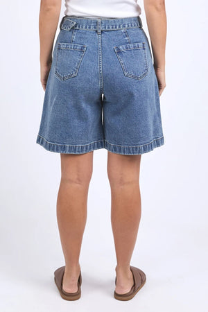 A woman faces away, wearing high-waisted Foxwood Saint Short in cotton with two back pockets, paired with a white top and brown sandals. The plain, light background highlights the Foxwood Saint Short as the outfitβs focal point.