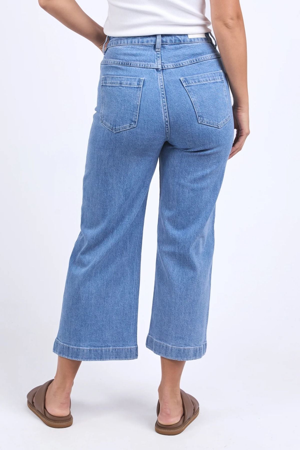 A person wearing the Foxwood Sarah Crop Jean in light blue, high-rise, wide-leg stretchy denim and brown sandals stands with their back to the camera against a plain white background.