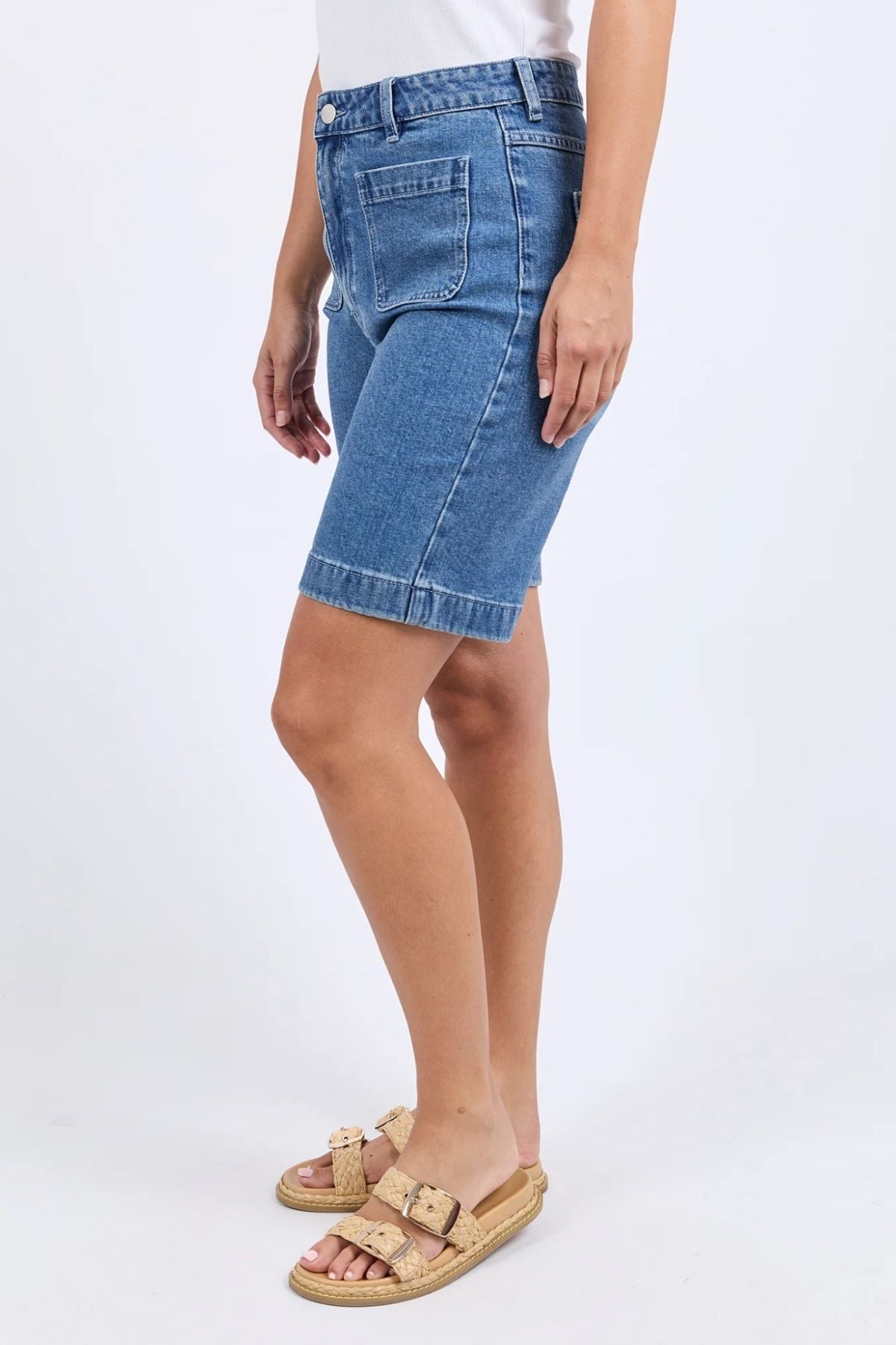 Someone stands against a plain light background, dressed in a white top, the knee-length blue denim Foxwood Sarah Short, and beige cork-sole sandals.