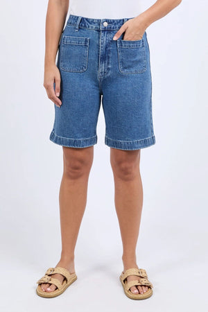 Wearing Foxwood Sarah Short denim Bermuda shorts with front patch pockets, a white top, and tan sandals, the person stands against a plain light background.