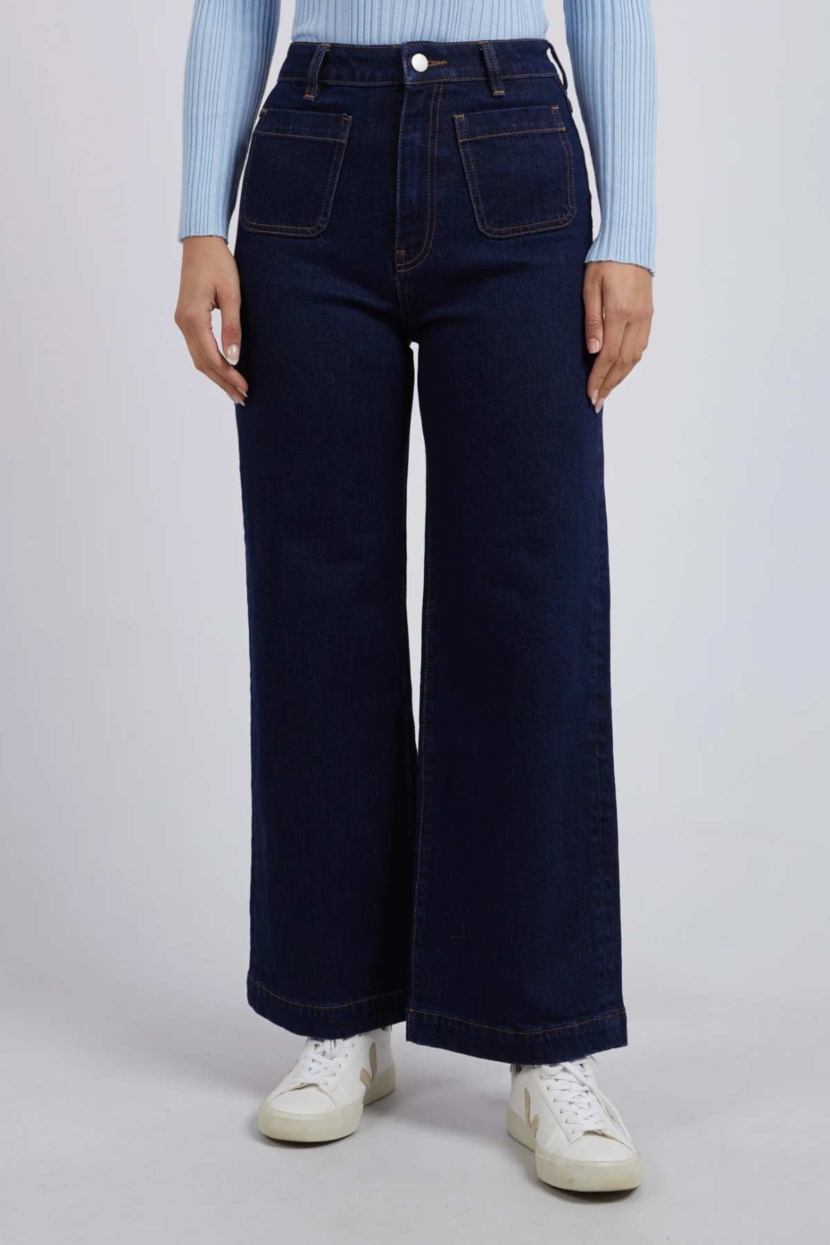 A person stands against a plain background wearing the Foxwood Sarah Wide Leg Jean in dark blue, made from a comfortable cotton elastane blend and featuring front patch pockets. The individual also wears a light blue long-sleeve top and white sneakers. The upper body is not visible in the image.