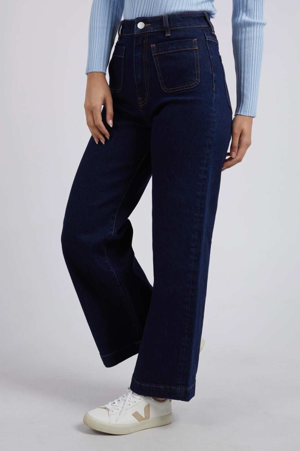 A person is seen from the waist down, wearing Foxwood Sarah Wide Leg Jeans in dark blue with prominent stitching, made of cotton elastane. They are paired with a light blue ribbed top and white sneakers against a plain, light background.