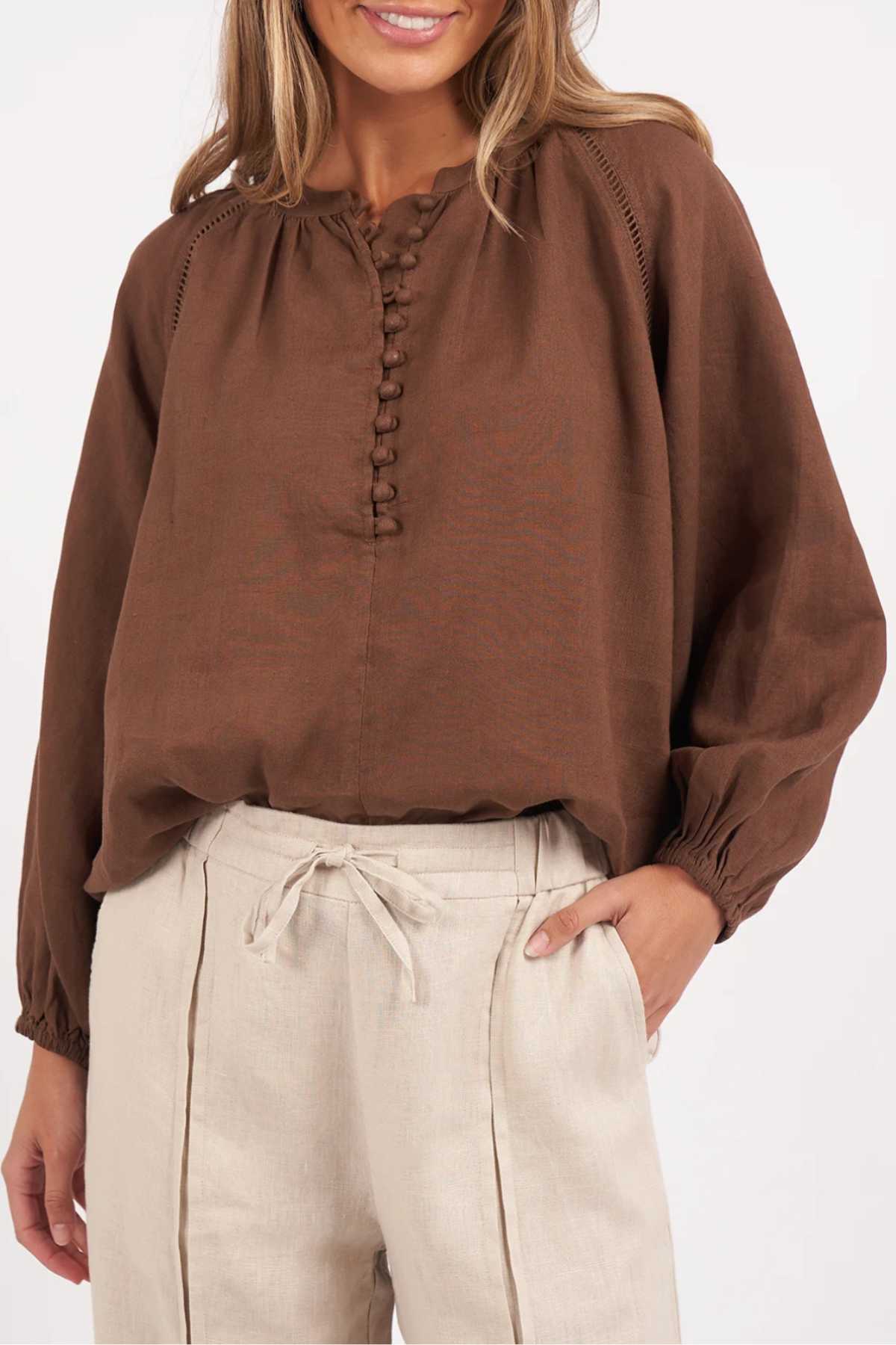A woman smiles with one hand in her pocket, wearing the Foxwood Sardinia Blouse—a loose brown top with fabric buttons and elastic balloon sleeves—paired with high-waisted, light beige drawstring pants.