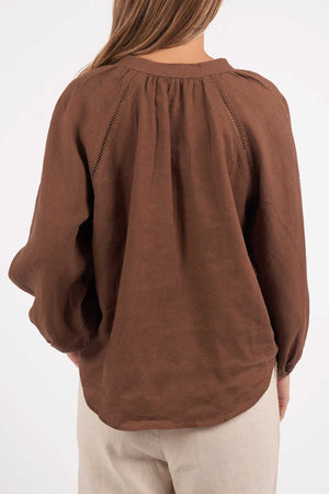 A woman stands with her back to the camera, highlighting the Foxwood Sardinia Blouse—a loose, long-sleeved brown top with gathered details and fabric-covered buttons—paired with light pants. Focus is on her upper body and the blouse's back.