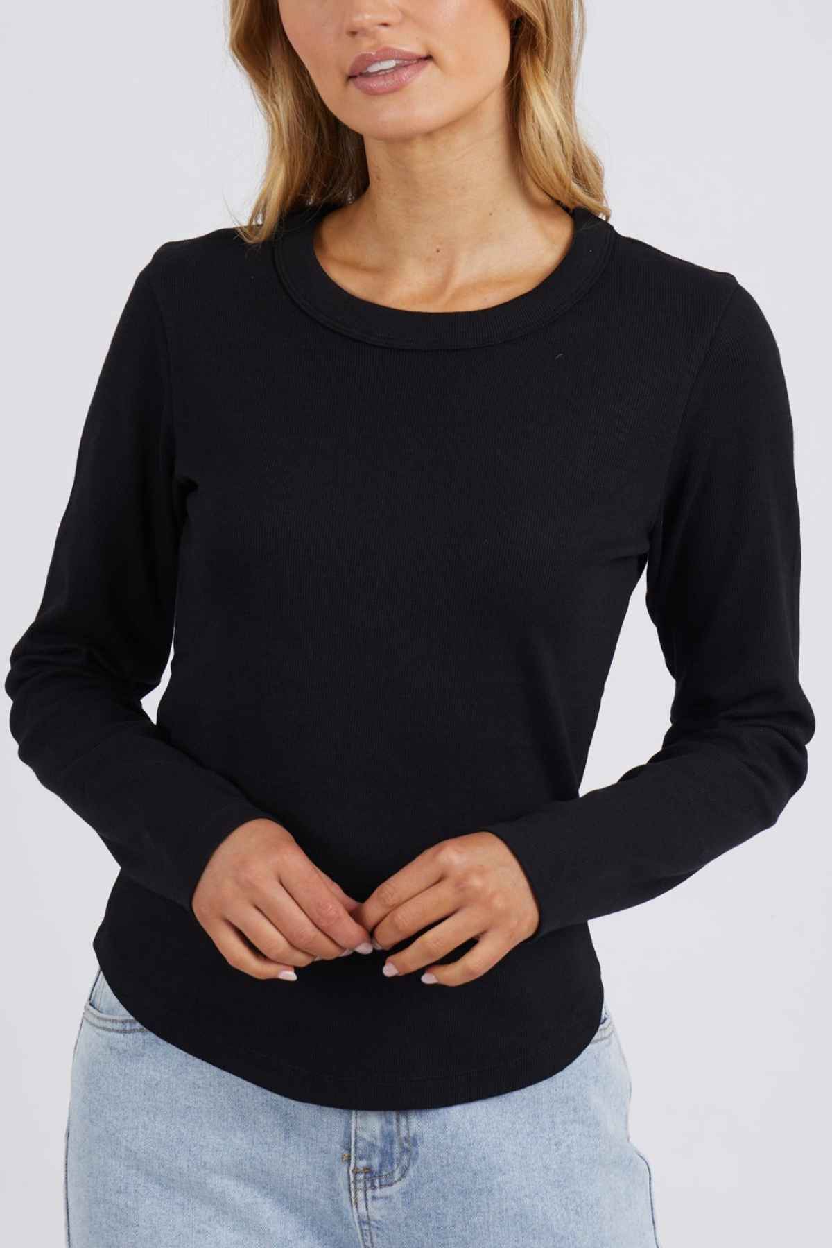 A woman in a Foxwood Scoop Long Sleeve Rib, black, paired with light blue jeans stands against a plain background. She glances slightly left, hands gently clasped, and her hair cascades loosely over her shoulders.