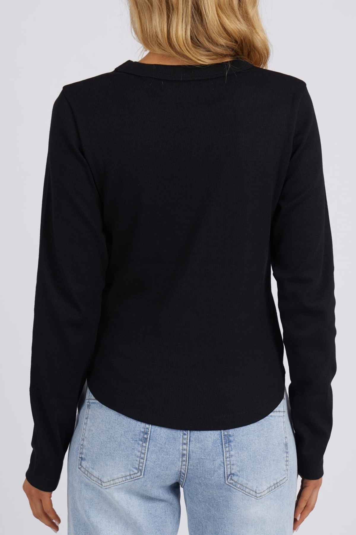 Rear view of a person with long blonde hair, wearing a Foxwood Scoop Long Sleeve Rib in black and light blue jeans, standing gracefully against a plain gray backdrop.