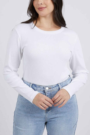 A person with long dark hair, wearing a plain white Foxwood Scoop Long Sleeve Rib shirt tucked into light blue high-waisted jeans, is standing against a plain background. The Foxwood scoop neckline adds charm as they smile gently, hands resting at their waist.