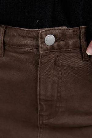 Close-up of a person wearing the Foxwood Scout Midi Skirt in brown denim with a silver button and visible stitching. One hand holds the waistband near the button, and a black top is tucked in.