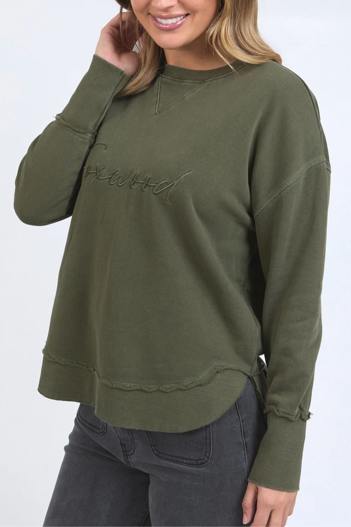 A woman smiles and touches her hair while wearing the Foxwood Signature Crew sweatshirt in olive green with subtle embroidered chest text, paired with gray jeans.