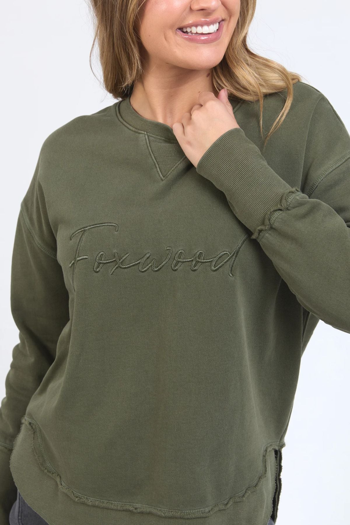 A woman smiles with one hand near her collar, wearing the Foxwood Signature Crew—a 100% cotton olive green crewneck sweater featuring "Foxwood" embroidered across the chest.