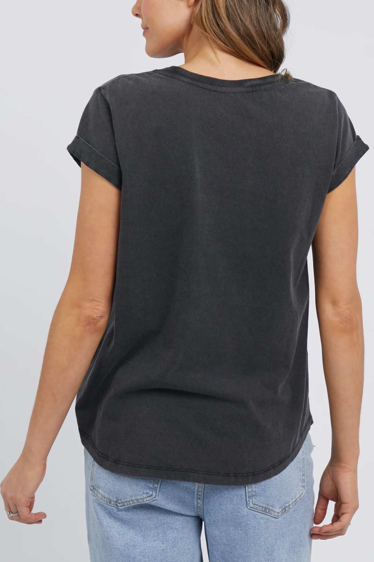 A woman is shown from the back, wearing the Foxwood Signature Tee in dark gray, with short sleeves that are slightly rolled. She has light brown hair and is paired with light blue denim jeans against a plain, light-colored background.