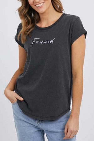 A person with long hair wears the Foxwood Signature Tee, a charcoal grey t-shirt featuring "Foxwood" written in cursive across the chest. With one hand in their pocket and dressed in light blue jeans, they smile comfortably, exuding a relaxed posture.