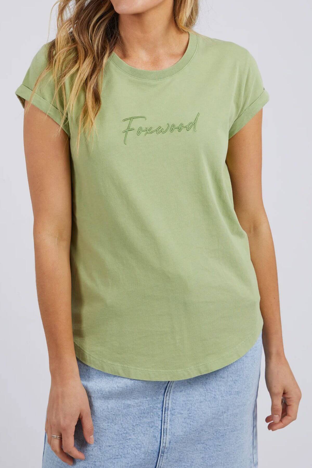 A person is wearing a Foxwood Signature Tee, light green, 100% cotton for breathable comfort, paired with a light denim skirt. The tee has rolled-up sleeves and appears against a plain white background, with the face not visible.