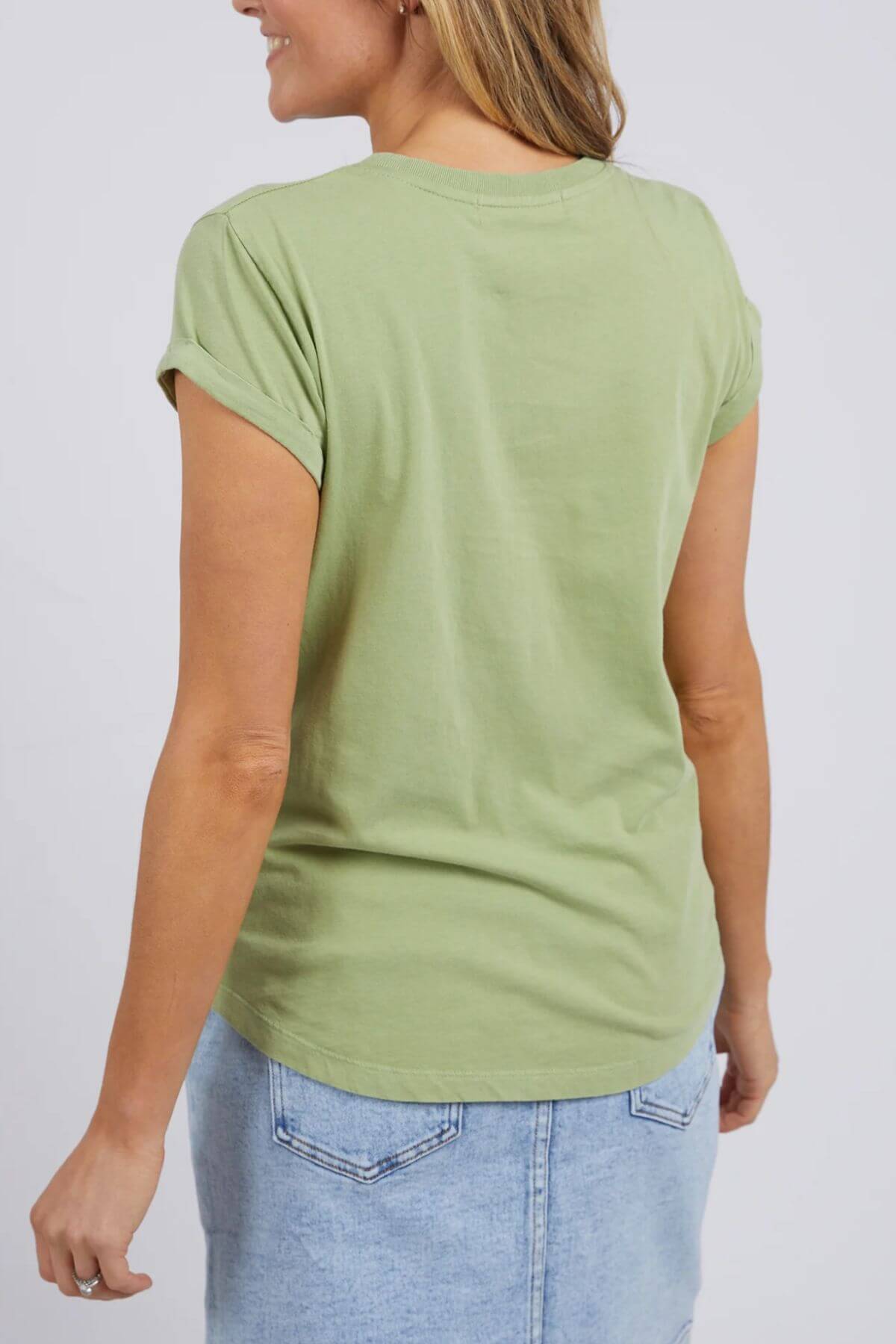 A person with light brown hair is seen from the back, wearing a 100% cotton Foxwood Signature Tee in light green paired with a light blue denim skirt, enjoying its lightweight, breathable fabric against a gray background.