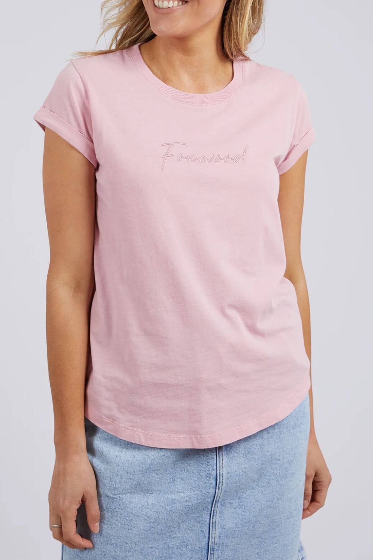 A woman smiles in the Foxwood Signature Tee, a 100% cotton light pink tee with rolled sleeves and subtle "Foxwood" text, paired with a light blue denim skirt.