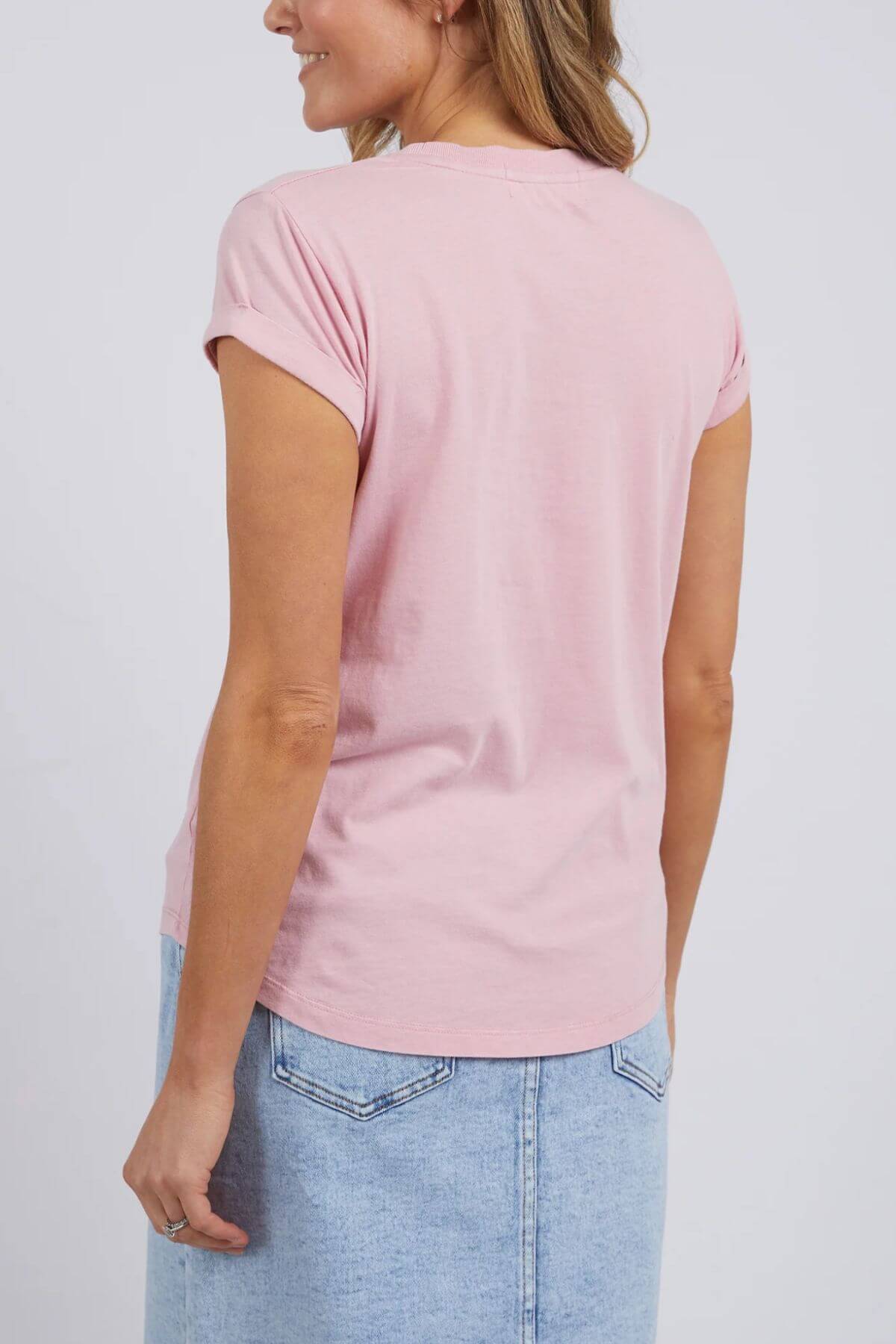 A woman with light brown hair is seen from behind wearing a light pink, 100% cotton Foxwood Signature Tee with a light blue denim skirt, standing against a plain background.