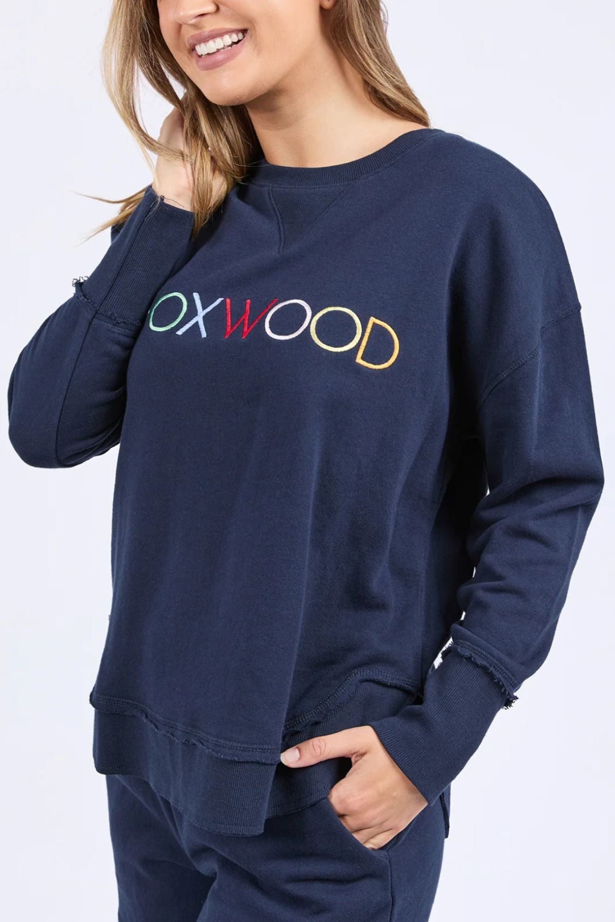 A woman smiles in the Foxwood Simplified Confetti Crew, a navy sweatshirt with multicoloured "OXWOOD" embroidery. Made from 100% cotton, she touches her hair with one hand and keeps the other in her pocket.