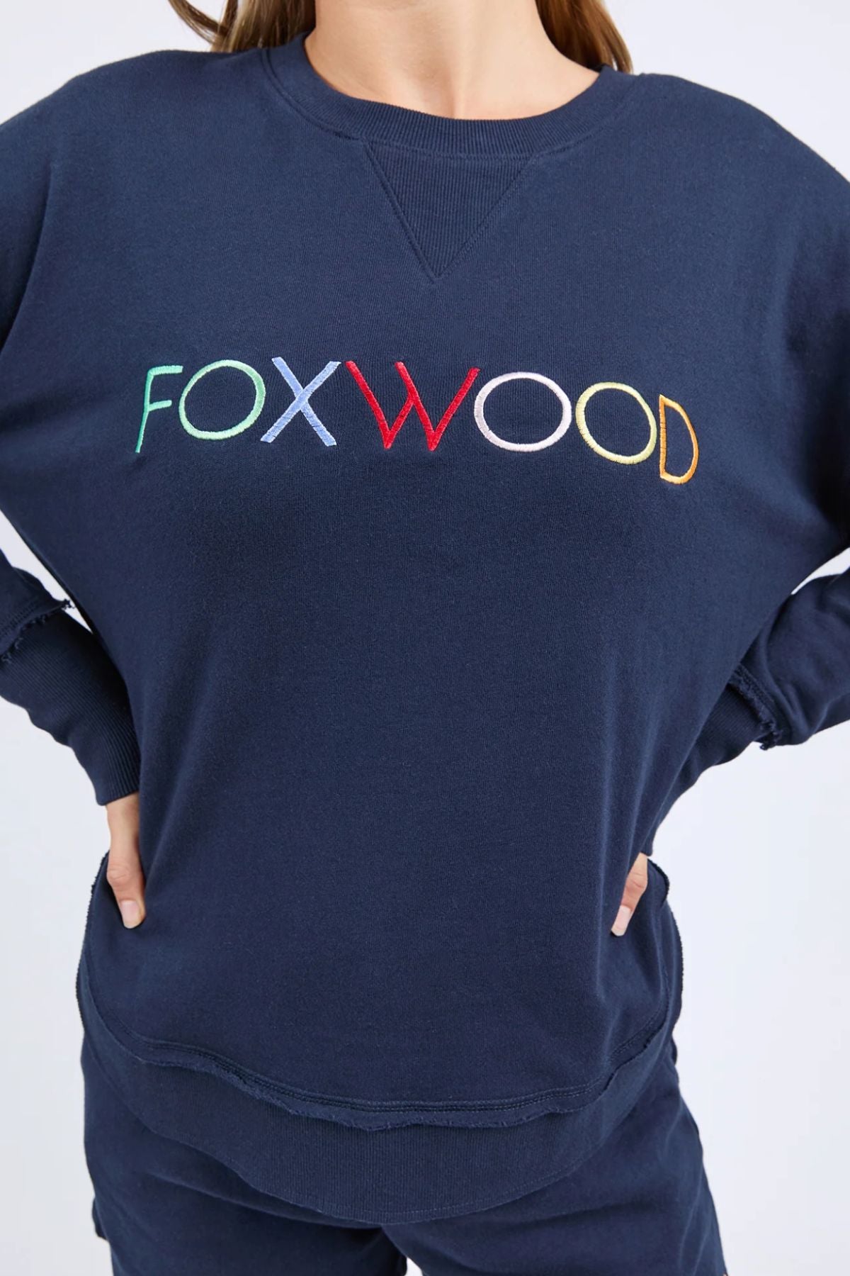 Wearing the Foxwood Simplified Confetti Crew, a navy blue 100% cotton sweatshirt featuring multicoloured "FOXWOOD" embroidery, their hands rest on their hips.