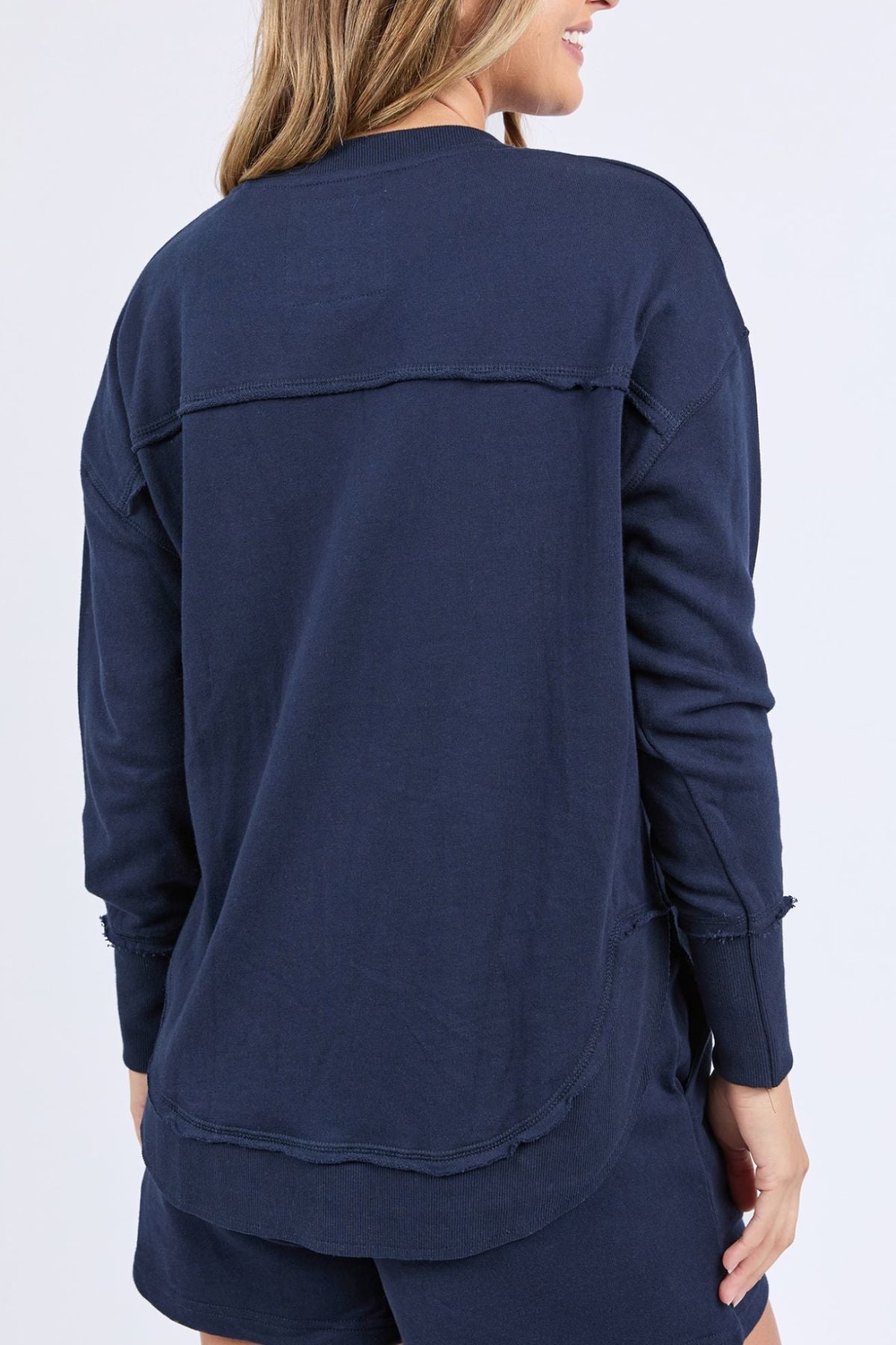 A woman stands with her back to the camera, wearing a Foxwood Simplified Confetti Crew sweatshirt with visible seam details and a relaxed fit, paired with matching navy blue shorts.