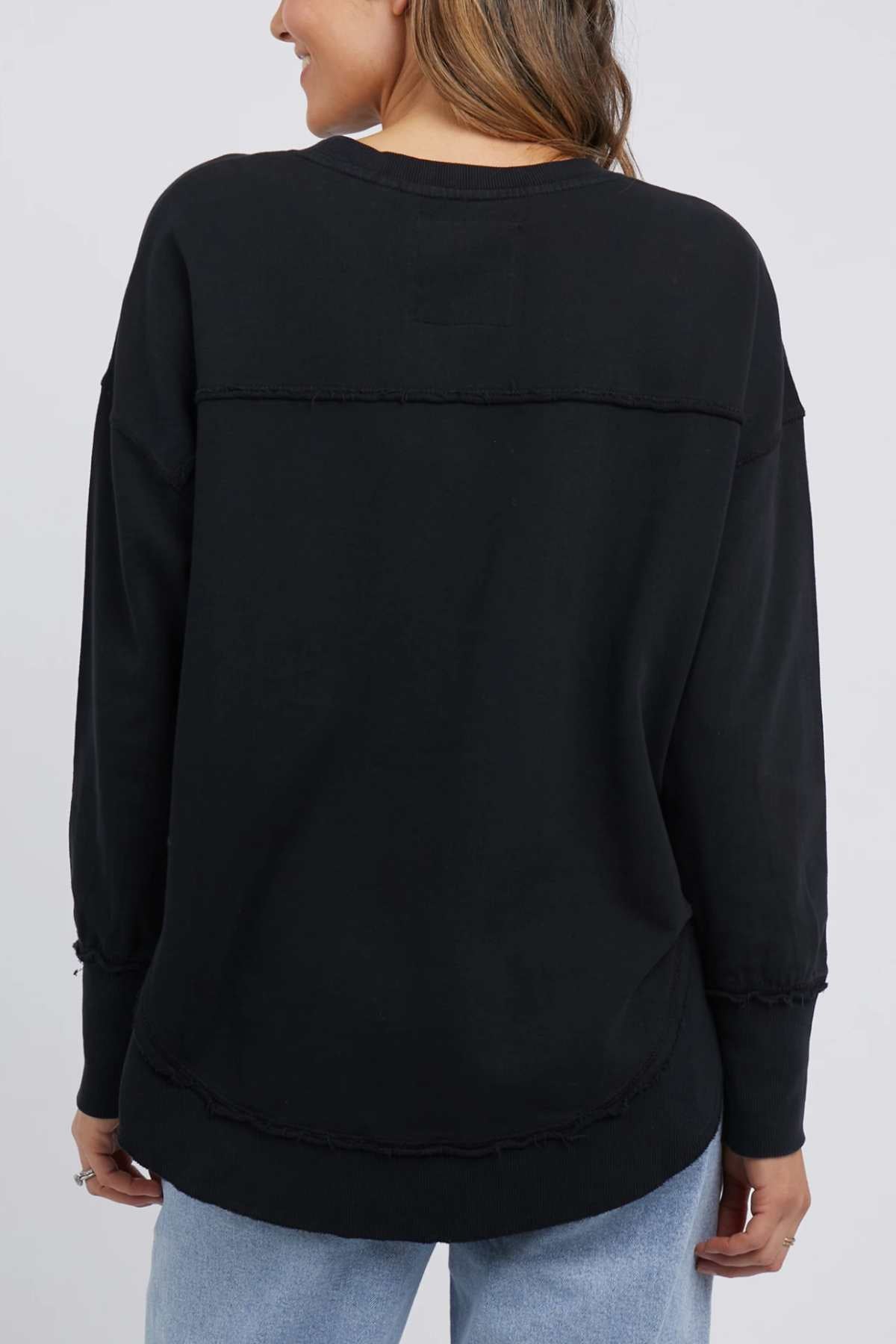 A person with long hair is seen from the back, dressed in a black Foxwood Simplified Crew and light blue jeans. The 100% cotton shirt features visible stitching details around the shoulders, waist, and sleeves. The background is plain and light-colored.