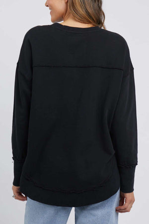 A person with long hair is seen from the back, dressed in a black Foxwood Simplified Crew and light blue jeans. The 100% cotton shirt features visible stitching details around the shoulders, waist, and sleeves. The background is plain and light-colored.