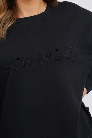 A person wearing a Foxwood Simplified Crew sweatshirt made of 100% cotton, featuring "FOXWOOD" embroidered across the chest in large letters. The image is cropped to highlight the detailed design of the sweatshirt and show part of the person's upper body.